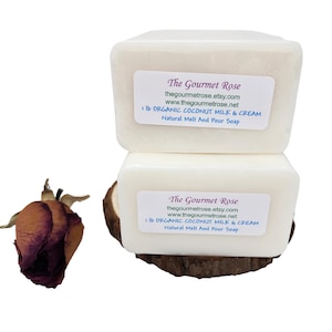 Op de afbeelding: Twee witte rechthoekige zeepblokken gestapeld op een houten basis, elk met een label met de tekst "The Gourmet Rose" en "1 lb ORGANIC COCONUT MILK & CREAM". Links een gedroogde roos. De zeep is voor natuurlijk smelten en gieten.