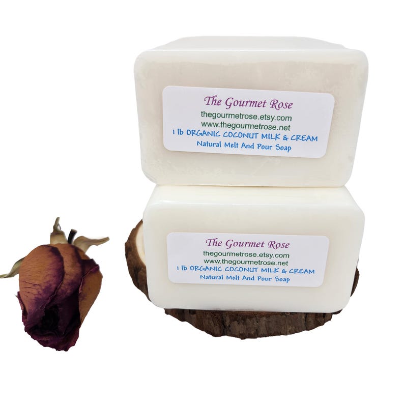 Op de afbeelding: Twee witte rechthoekige zeepblokken gestapeld op een houten basis, elk met een label met de tekst "The Gourmet Rose" en "1 lb ORGANIC COCONUT MILK & CREAM". Links een gedroogde roos. De zeep is voor natuurlijk smelten en gieten.