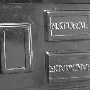 May include: A rectangular, dark gray mold with three recessed areas. The words "NATURAL" and "HANDMADE" are embossed in two of the rectangular areas. The mold is likely for soap or other craft items.