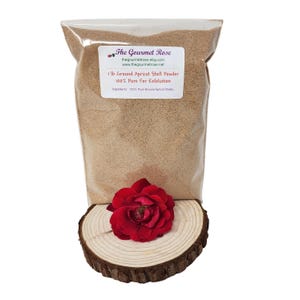 Op de afbeelding: Een doorzichtige zak gevuld met lichtbruin poeder, met het label "1 lb Ground Apricot Shell Powder 100% Pure For Exfoliation." Een helderrode roos ligt op een houten schijf. Het logo The Gourmet Rose is zichtbaar.