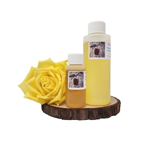 May include: Two bottles of liquid, one large and one small, with white caps and labels. A yellow rose and a wooden slice are also in the image. The bottles contain a light yellow and tan liquid. The labels have a brown and white design.