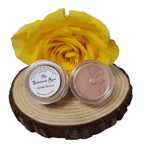 Può includere: Due piccoli vasetti trasparenti di trucco minerale sono su una fetta di legno, con una rosa gialla sullo sfondo. Un vasetto è etichettato "The Gourmet Rose Adobe Desert". Il trucco è di colore marrone chiaro.