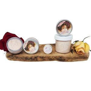 Könnte beinhalten: Eine Sammlung von Mineral-Foundation-Produkten von "The Gourmet Rose" auf einem Holzbrett. Die Produkte umfassen Gläser und Dosen mit Etiketten, die das Porträt einer Frau zeigen. Eine rote Rose und gelbe Rosen sind ebenfalls vorhanden.