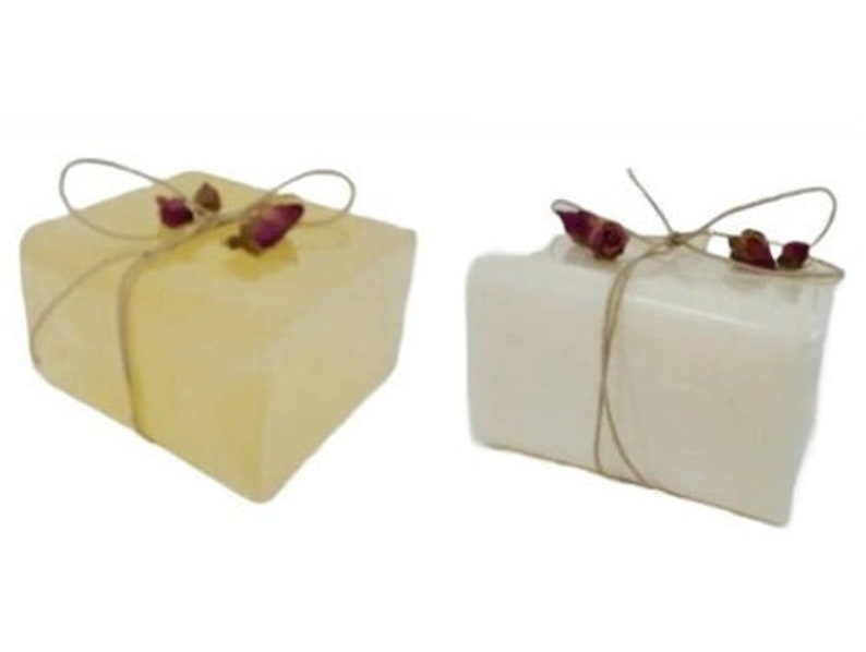 May include: Two square soaps, one pale yellow and one white, are tied with twine and adorned with dried rosebuds. The soaps have a rustic, handmade appearance, suggesting a natural skincare product.