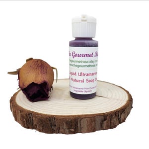 K&ouml;nnte beinhalten: Eine Flasche fl&uuml;ssiger ultramarinfarbener nat&uuml;rlicher Seifenfarbe mit wei&szlig;em Verschluss, neben einer getrockneten Rose auf einer Holzscheibe. Die Flasche hat ein Etikett mit dem Text "The Gourmet Rose" und "Liquid Ultramarine Natural Soap Dye".