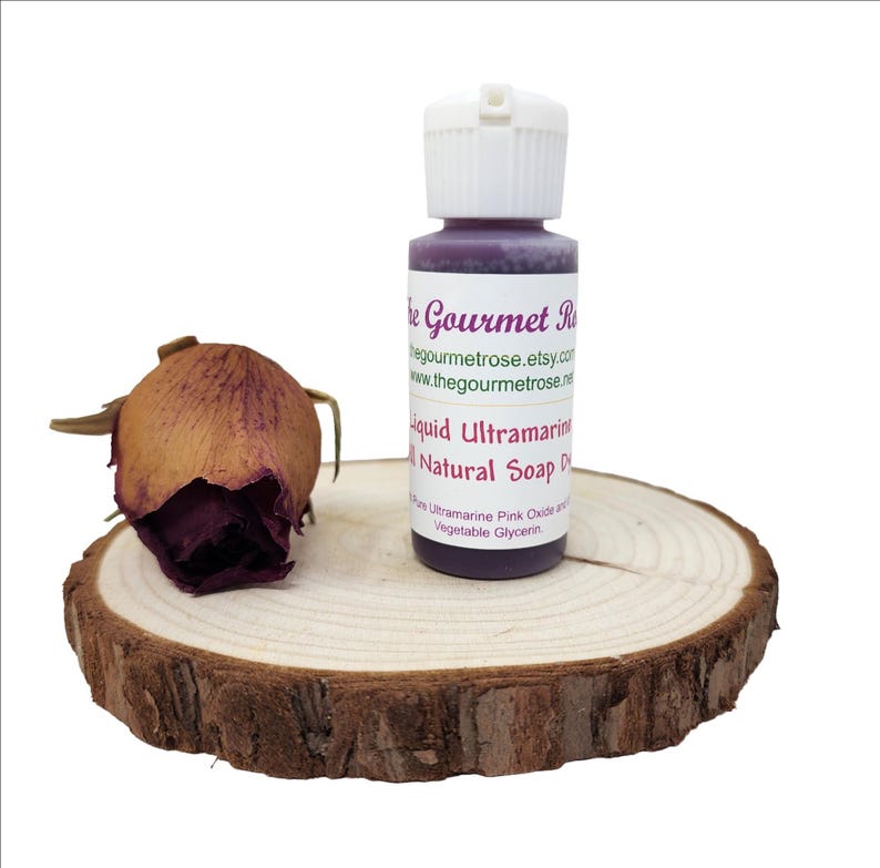 K&ouml;nnte beinhalten: Eine Flasche fl&uuml;ssiger ultramarinfarbener nat&uuml;rlicher Seifenfarbe mit wei&szlig;em Verschluss, neben einer getrockneten Rose auf einer Holzscheibe. Die Flasche hat ein Etikett mit dem Text "The Gourmet Rose" und "Liquid Ultramarine Natural Soap Dye".