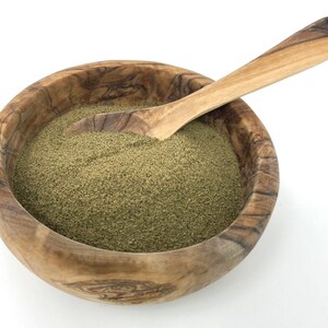 Puede incluir: Un cuenco de madera lleno de polvo verde, posiblemente una especia o hierba, con una cuchara de madera. El cuenco tiene un patr&oacute;n de vetas de madera natural y est&aacute; sobre un fondo blanco. La cuchara es de color marr&oacute;n claro y parece ser de la misma madera.