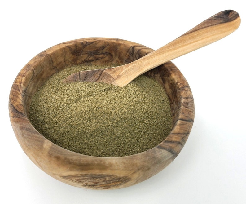 Puede incluir: Un cuenco de madera lleno de polvo verde, posiblemente una especia o hierba, con una cuchara de madera. El cuenco tiene un patr&oacute;n de vetas de madera natural y est&aacute; sobre un fondo blanco. La cuchara es de color marr&oacute;n claro y parece ser de la misma madera.