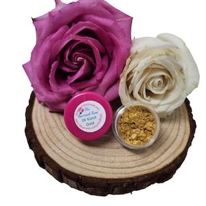 Pode incluir: Uma fatia de madeira com um recipiente rosa e um recipiente transparente com flocos de ouro. O recipiente rosa tem um rótulo branco com o texto "The Gourmet Rose 24 Karat Gold". Duas rosas, uma rosa e uma branca, no fundo.