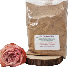 Puede incluir: Una bolsa de plástico transparente llena de polvo de cáscara de nuez molida, etiquetada como "The Gourmet Rose". Una rosa rosa y una rodaja de madera están en primer plano. La etiqueta dice "1 lb Ground Walnut Shell Powder 100% Pure For Exfoliation."