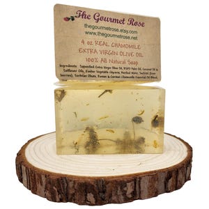 Könnte beinhalten: Ein klarer, rechteckiger Seifenblock mit sichtbaren Kamillenblüten und Kräutern, der auf einer Holzscheibe ruht. Die Seife ist mit "4 oz REAL CHAMOMILE EXTRA VIRGIN OLIVE OIL 100% All Natural Soap" und dem Markennamen "The Gourmet Rose" beschriftet.