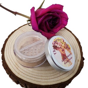 Può includere: Un barattolo di cipria abbronzante con la scritta "The Gourmet Rose GRAND MARNIER Bronzer Powder" sul coperchio. Una rosa rosa intenso poggia su una fetta di legno. L'abbronzante è di colore marrone chiaro.