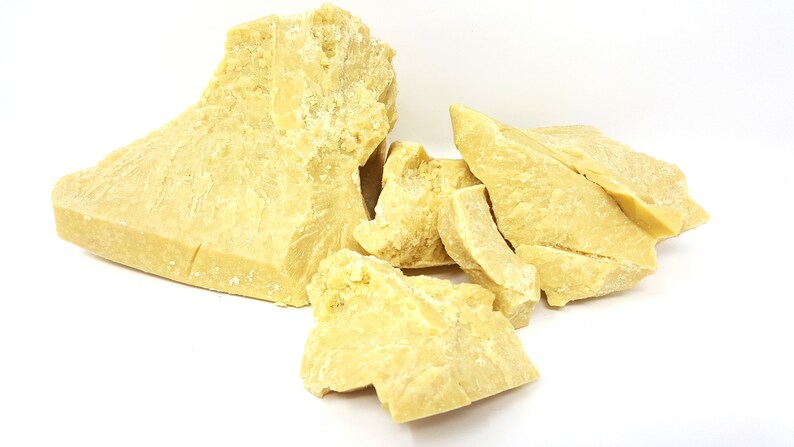 May include: Chunks of raw, unrefined cocoa butter, a pale yellow solid with a slightly rough texture. The pieces vary in size and shape, with a few large blocks and several smaller fragments. The background is plain white.