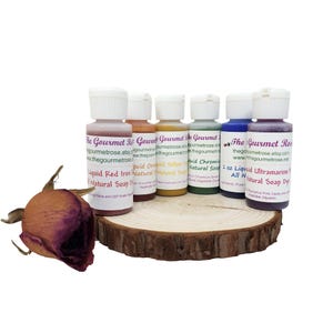 May include: A collection of small bottles of natural soap dye in various colors, including red, orange, yellow, green, blue, and purple. The bottles are arranged on a wooden slice, with a dried rose in the foreground. The labels read "Liquid Red Iron Natural Soap", "Liquid Orchid Yellow Natural Soap", and "Liquid Ultramarine Natural Soap Dye".