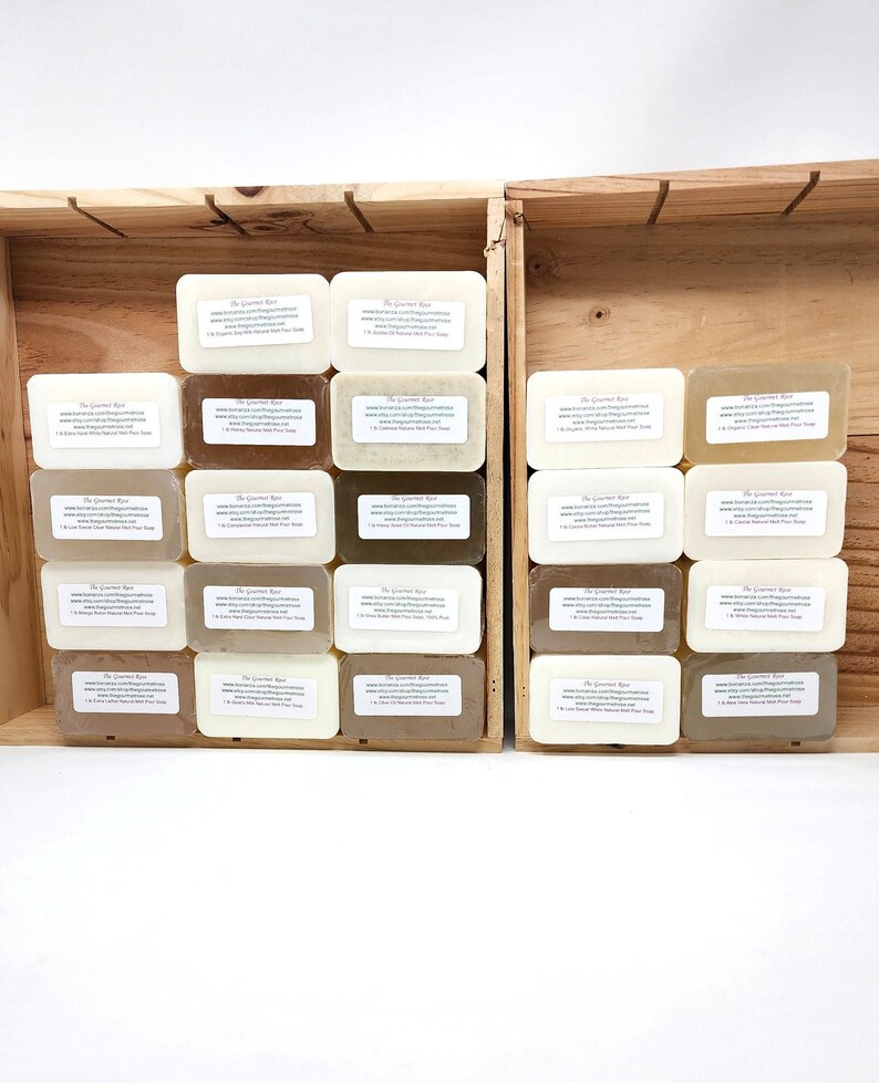 Puede incluir: Barras rectangulares de jab&oacute;n natural en varios tonos de marr&oacute;n, beige y blanco, dispuestas dentro de una caja de madera. Cada barra tiene una etiqueta con texto. La caja est&aacute; abierta, mostrando los jabones, sobre una superficie blanca.