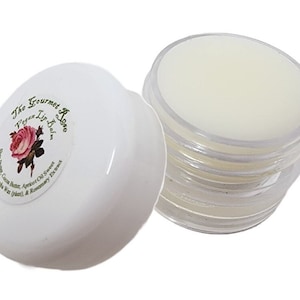 May include: A white lip balm container with a clear, open container next to it. The white container has a label with the text "The Gourmet Rose Vegan Lip Balm" and a rose illustration. The open container shows the balm.