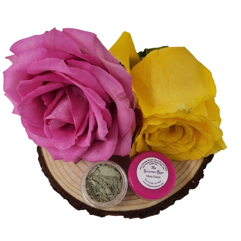 Puede incluir: Una composici&oacute;n con una rosa rosa vibrante y una rosa amarilla, un frasco transparente de polvo verde y un frasco rosa etiquetado "Moss Green". Los elementos est&aacute;n dispuestos sobre una rodaja de madera.