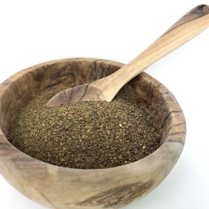 Puede incluir: Un cuenco de madera lleno de pimienta negra molida gruesa, con una cuchara de madera dentro. El cuenco y la cuchara son de madera de color claro, y la pimienta es de color verde oscuro y terroso.