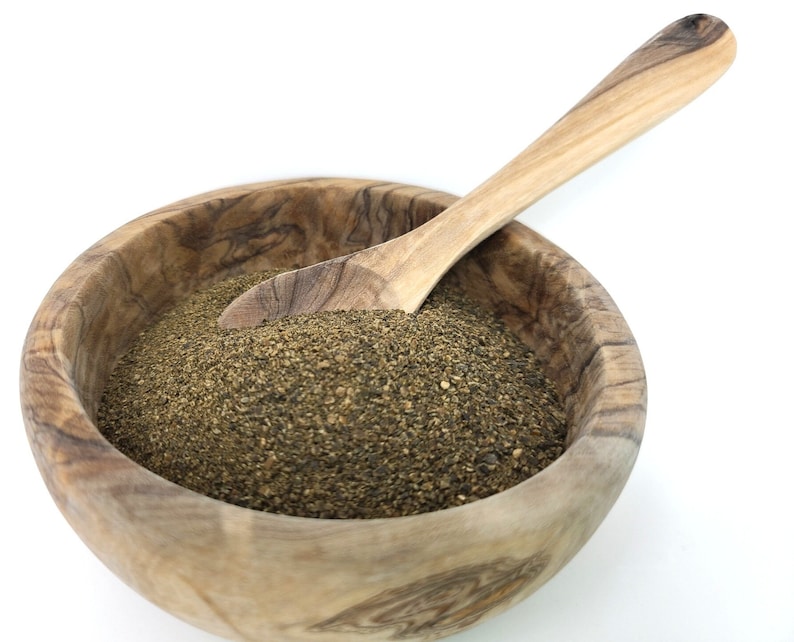 Puede incluir: Un cuenco de madera lleno de pimienta negra molida gruesa, con una cuchara de madera dentro. El cuenco y la cuchara son de madera de color claro, y la pimienta es de color verde oscuro y terroso.