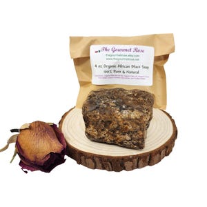 May include: A brown, rectangular block of organic African black soap, labeled as 4 oz, sits on a wooden slice. A dried rose and a paper bag with text are also in the image. The text on the bag reads "The Gourmet Rose" and "100% Pure & Natural."