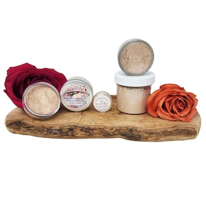 May include: A wooden serving board displays several jars of face powder and two roses. The jars are filled with light brown powder. One rose is deep red, and the other is orange. The text on the jars reads "Fair Light Concealer" and "The Gourmet Barn".