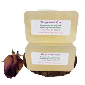 May include: Two rectangular bars of clear, organic soap stacked on a wooden base. Each bar has a white label with the text "The Gourmet Rose" and product information. A dried rose bud is visible on the left side.