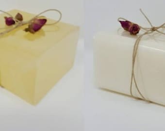 Puede incluir: Dos barras de jabón, una amarilla y otra blanca, están envueltas en celofán transparente y atadas con hilo. Cada barra está adornada con capullos de rosa secos. El jabón tiene un aspecto natural y artesanal.
