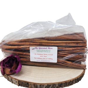 May include: A clear plastic bag filled with cinnamon sticks, approximately 10 inches long, sits on a wooden slice. A label reads "1 lb Cinnamon Sticks, 10 Inch 100% Pure For Culinary Use & Crafting." A dried purple rose is on the left.