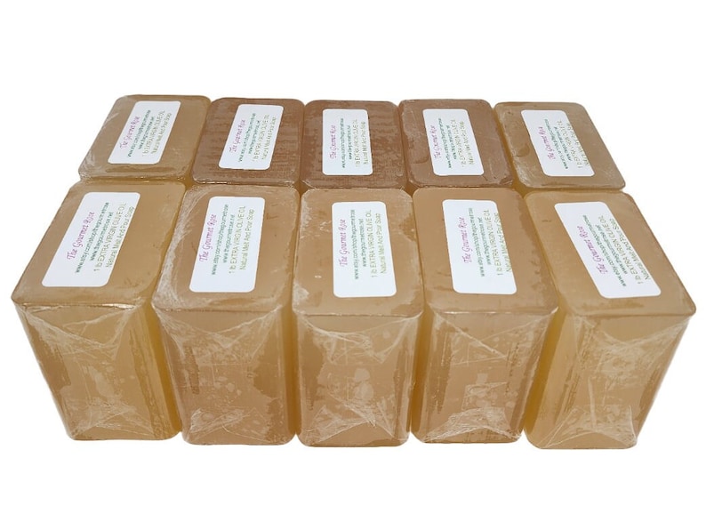 May include: A collection of rectangular, light amber-colored soaps, individually wrapped in clear plastic. Each soap has a white label with text. The soaps are arranged in a neat, stacked formation, showcasing a natural, handcrafted aesthetic.