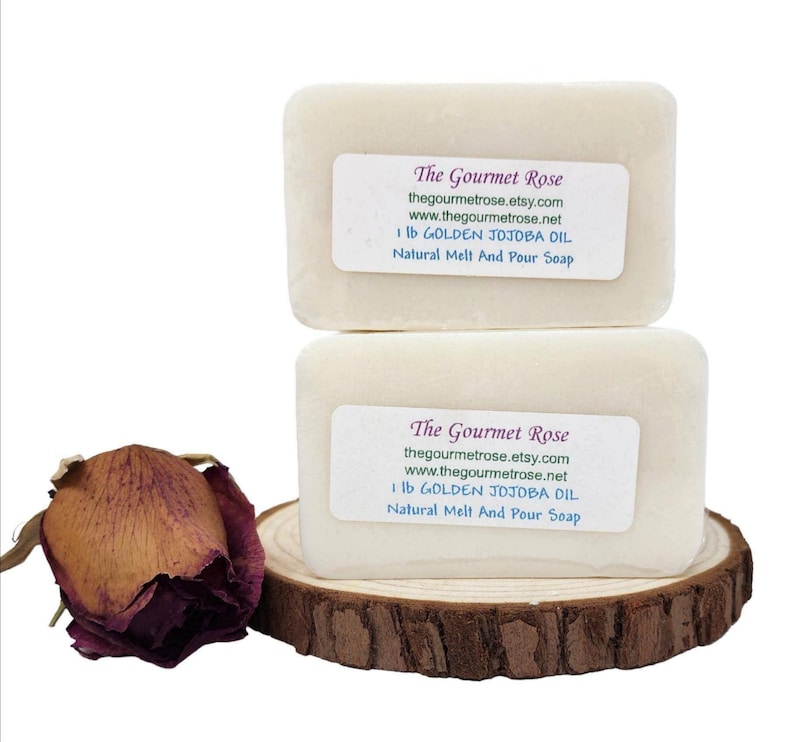 Op de afbeelding: Twee rechthoekige blokken natuurlijke smelt- en gietzeep, met het label "The Gourmet Rose" met website-informatie en "1 lb GOLDEN JOJOBA OIL". De zeep ligt op een houten schijf, met een gedroogde rozenknop.