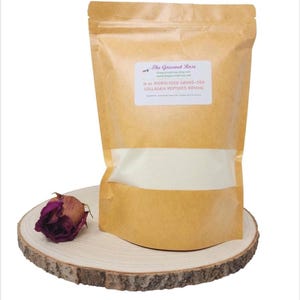 May include: A brown paper bag of collagen peptides with a clear window, labeled "The Gourmet Rose." A dried, dark purple rose sits on a wooden slice next to the bag. The label reads "6 oz Hydrolyzed Grass-Fed Collagen Peptides Bovine."