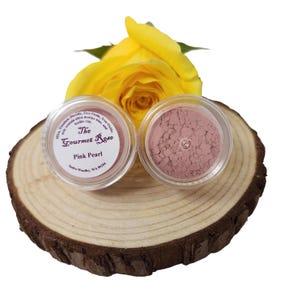 Può includere: Due piccoli vasetti trasparenti di cipria cosmetica rosa perla sono su una fetta di legno, con una rosa gialla sullo sfondo. Un vasetto ha un'etichetta con scritto "The Gourmet Rose Pink Pearl". L'altro vasetto è aperto, rivelando la cipria rosa.