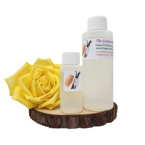 Puede incluir: Dos botellas blancas de aceite perfumado, una grande y otra pequeña, sobre una rodaja de madera. A la izquierda, una rosa amarilla. Las etiquetas dicen "Vainilla Cálida" e incluyen una ilustración de vainilla.