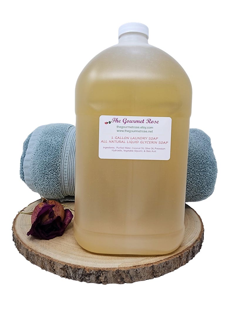 Peut inclure: Un grand bidon en plastique transparent de savon &agrave; lessive avec un bouchon blanc, &eacute;tiquet&eacute; "1 GALLON LAUNDRY SOAP". Le bidon est pos&eacute; sur une tranche de bois avec une rose s&eacute;ch&eacute;e et deux serviettes bleues. L'&eacute;tiquette comprend l'adresse du site web.