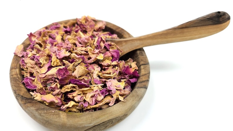 May include: A wooden bowl filled with dried rose petals, in shades of pink and tan, sits next to a wooden spoon. The bowl and spoon are made of light-colored wood, and the petals are scattered throughout.