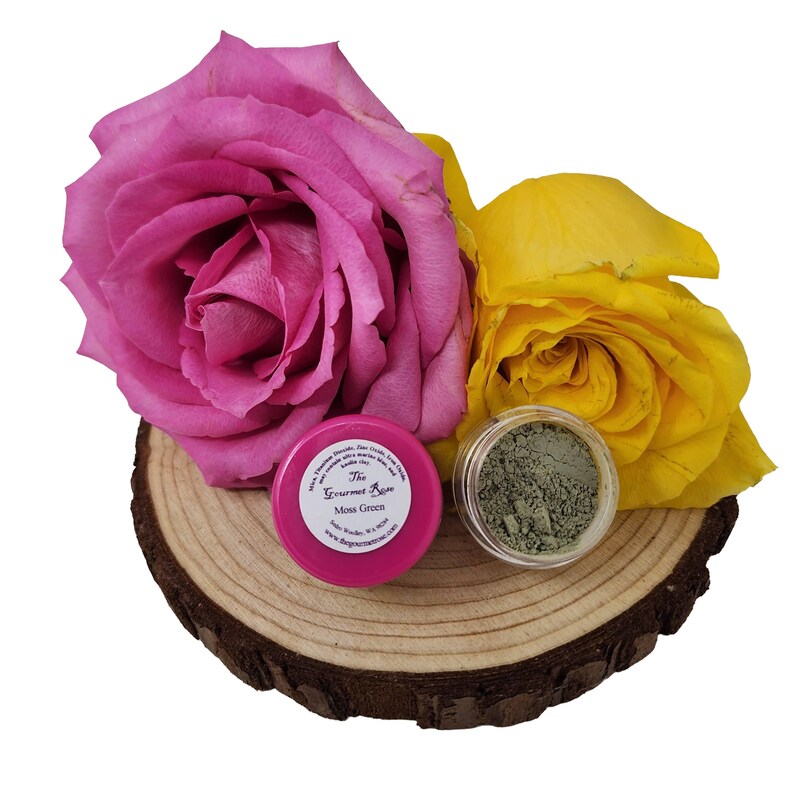 Puede incluir: Un frasco rosa etiquetado "The Gourmet Rose Moss Green" y un frasco transparente de polvo verde sobre una rodaja de madera. Una gran rosa rosa y una rosa amarilla en el fondo. Los frascos est&aacute;n sobre una rodaja de madera.