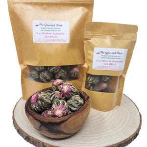 May include: Organic blooming tea balls in a wooden bowl and two kraft paper bags. The bags are labeled with the text "The Gourmet Rose" and "Organic Blooming Tea Balls". The bowl sits on a wooden slice.