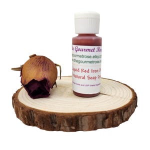 May include: A small bottle of red liquid soap dye with a white cap is on a wooden slice next to a dried rose bud. The label reads "Liquid Red Iron Oxide Natural Soap Dye". The Gourmet Rose Etsy shop is listed.