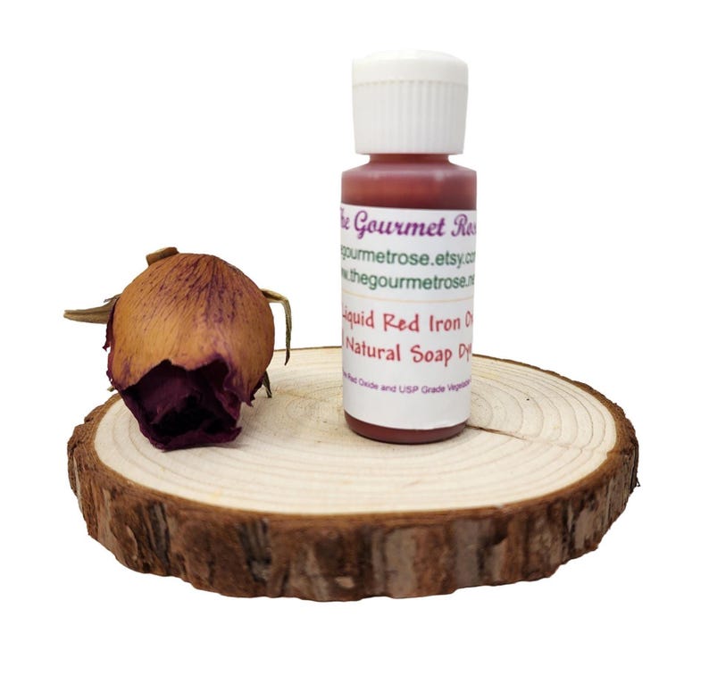 May include: A small bottle of red liquid soap dye with a white cap sits on a wooden slice next to a dried rose bud. The label reads "Liquid Red Iron Oxide Natural Soap Dye". The Gourmet Rose Etsy shop is also listed.
