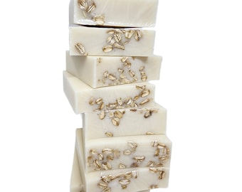 May include: A stack of rectangular, off-white soap bars with visible embedded oatmeal flakes. The bars are arranged in a vertical tower, showcasing the texture and natural ingredients of the soap. The background is plain white.