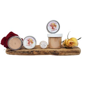 Op de afbeelding: Een display van mineral veil make-up producten op een houten plank, waaronder potten en blikken van verschillende formaten. Een rode roos en een gele roos zijn aan weerszijden van de producten geplaatst. De labels bevatten een vintage illustratie.