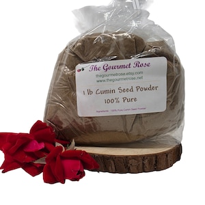 Può includere: Un sacchetto di plastica trasparente pieno di polvere di semi di cumino, etichettato "1 lb Cumin Seed Powder 100% Pure". Il sacchetto poggia su una fetta di legno, con rose rosse in primo piano. L'etichetta include il sito web thegourmetrose.etsy.com.