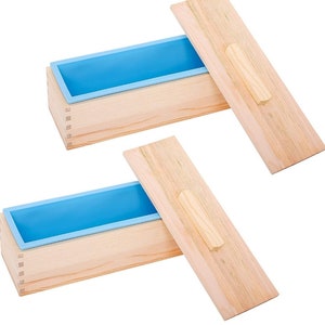 Può includere: Due stampi per sapone in legno con rivestimenti in silicone blu e coperchi in legno. Gli stampi rettangolari sono realizzati in legno chiaro con giunti a coda di rondine. I coperchi hanno un manico in legno.