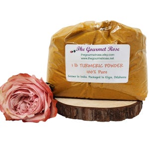 Puede incluir: Una bolsa transparente llena de polvo de cúrcuma amarillo brillante, etiquetada como "1 lb TURMERIC POWDER 100% Pure". Una rosa rosa y una rodaja de madera están en primer plano. La etiqueta también incluye la dirección del sitio web.