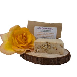 May include: A bar of oatmeal, milk, and honey soap with a label from The Gourmet Rose, resting on a wooden slice. A yellow rose is to the left. The soap is decorated with oatmeal flakes.