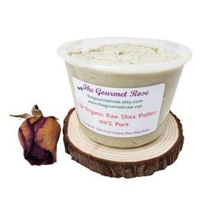 May include: A clear plastic container filled with off-white shea butter, labeled "The Gourmet Rose." The label indicates it contains 1 lb of organic raw shea butter, 100% pure. The container sits on a small, round wooden coaster, with a dried rose bud to the left.