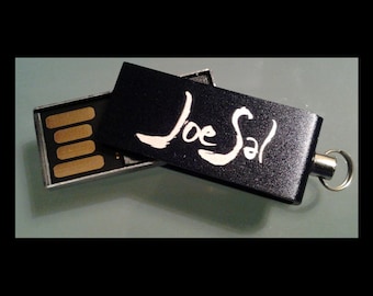 May include: A black USB flash drive with a silver metal casing. The drive has a silver metal loop for attaching a keychain. The drive is open and the inscription "Joe Sal" is visible on the black plastic cover.
