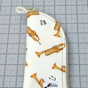 May include: A white zippered pouch with a curved shape. The pouch has a gold zipper and a white background with a repeating pattern of gold trumpets and musical notes. The pouch has a red and white label with the word "Betta" printed on it.