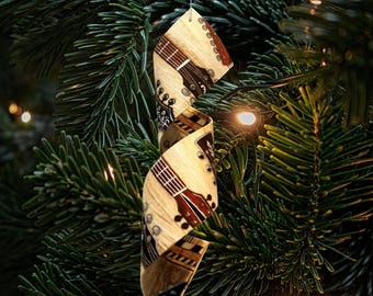 Puede incluir: Adorno decorativo para árbol de Navidad con forma de cinta, con un patrón repetido de guitarras acústicas. El adorno es de madera clara con detalles de guitarra marrones y rojos, sobre un fondo de agujas de pino verdes.