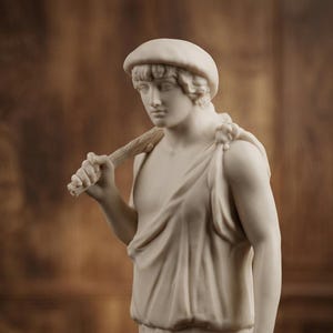 Op de afbeelding: Een klassieke sculptuur van een jonge persoon, gebeeldhouwd in een crèmewitte steen. De figuur draagt een pet en gedrapeerde kleding, met een houten staf over de schouder. Het beeld heeft een serene uitdrukking en staat tegen een vage bruine achtergrond.